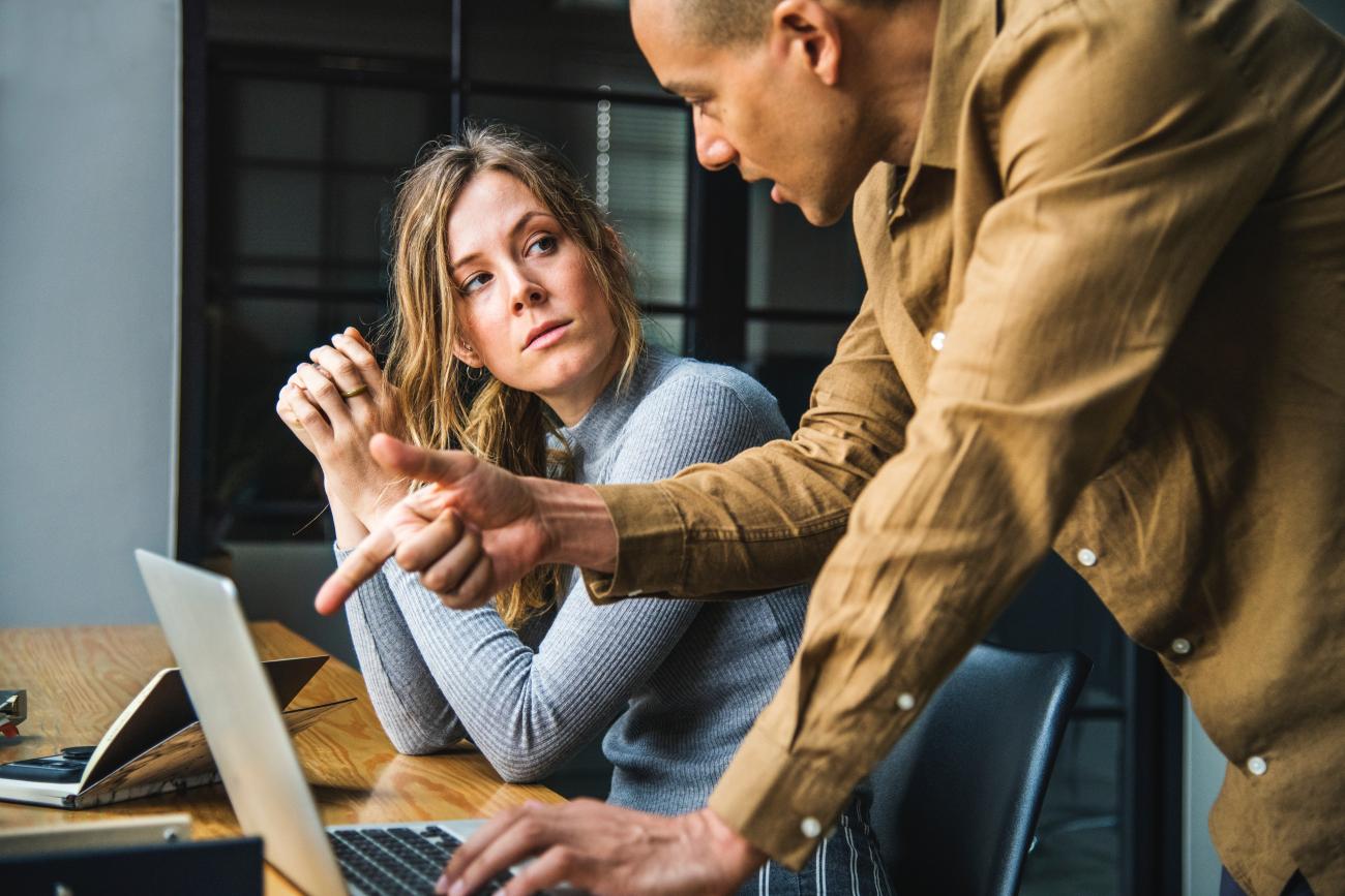 Image of a man and woman looking at a laptop screen and having a discussion about it. She is wearing a blue top and is listening to him, he is wearing a brown shirt and pointing at the screen whilst talking.