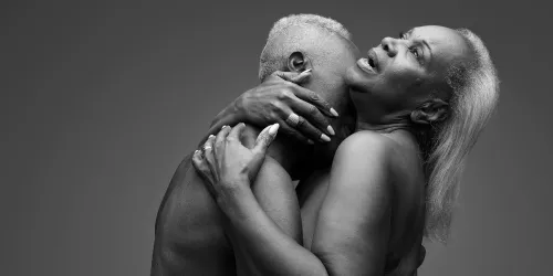 An elderly couple embrace, he kisses her neck while she stares off in ecstasy